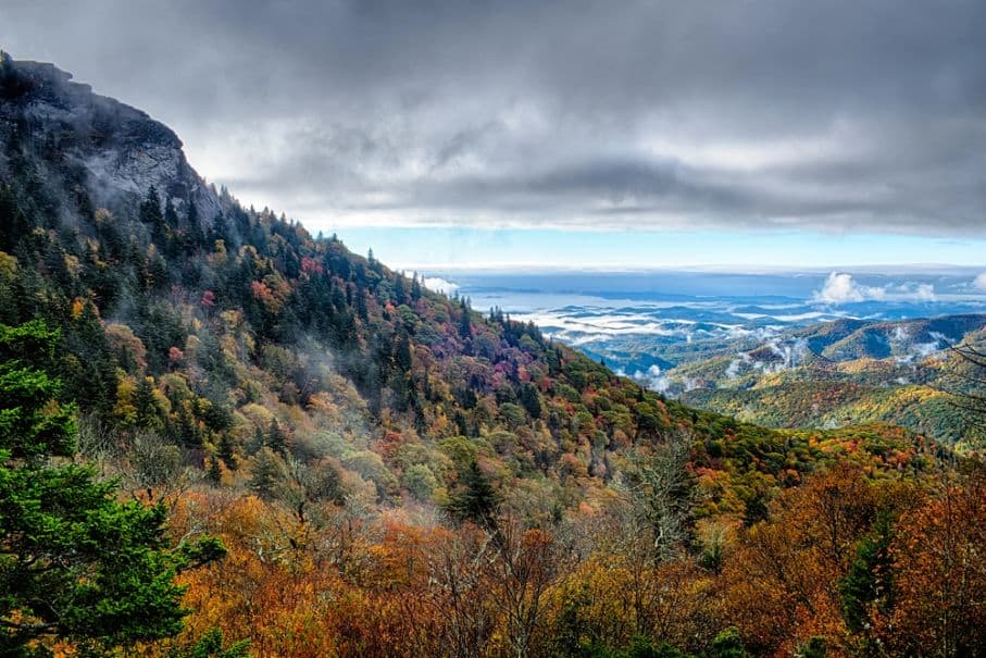 Mountain landscape with colorful autumn trees, fog, and low clouds. A rocky peak rises on the left, while valleys, with cabins near Fairy Stone State Park tucked away in their embrace, extend into the distance under a cloudy sky.